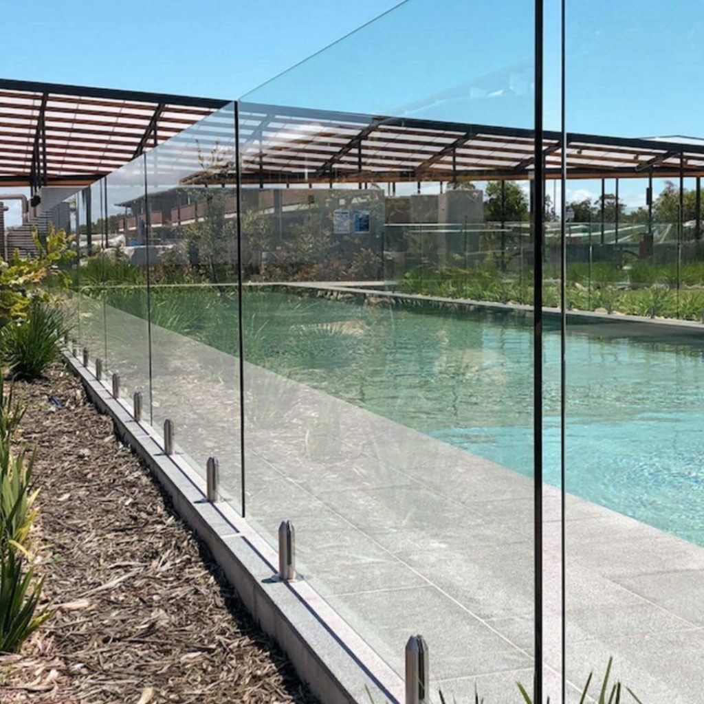 A clear and modern glass pool fence encloses a swimming pool, providing a safe and unobtrusive barrier. The glass panels are secured with sleek stainless steel mounts along the edge of the pool area.