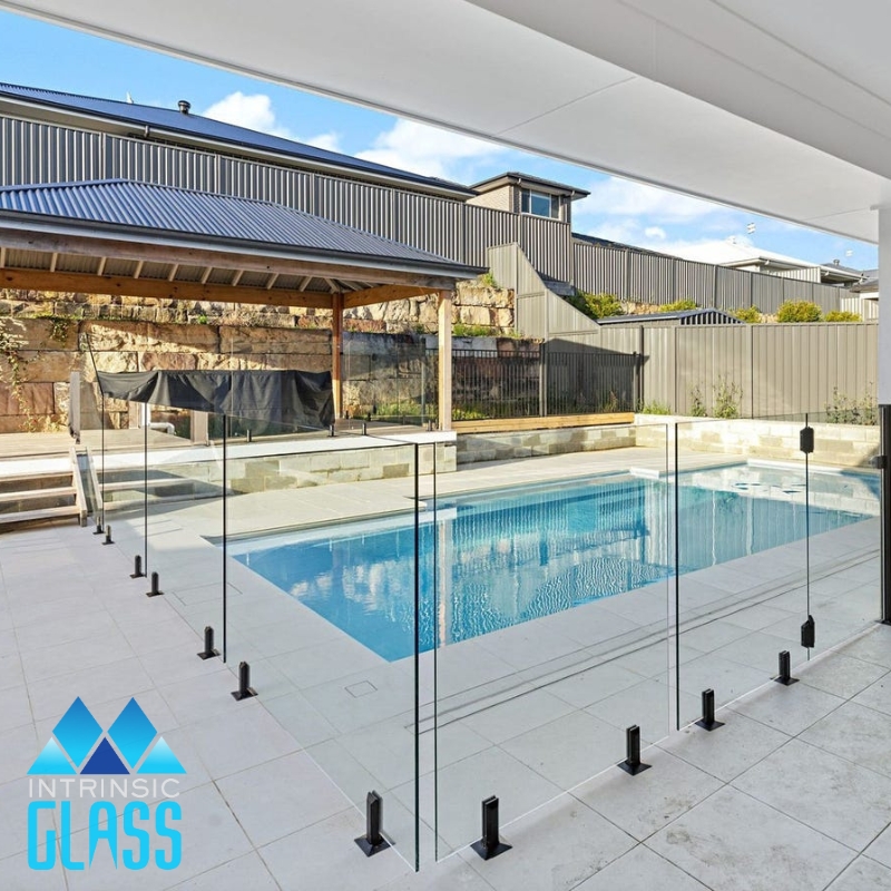 A modern swimming pool with a transparent glass pool fencing Blacksmiths. The fence has a black frame and is installed around the perimeter of the pool. In the background, there is a patio with a grill.