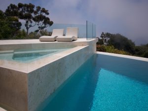 Elegant infinity pool with modern glass pool fence, luxurious loungers, marble deck, and a scenic view of nature.