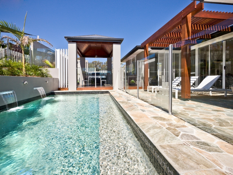 Luxury outdoor area with modern glass pool fence, sleek pool, wooden pergola, lounge chairs, and water features.