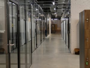 Modern office hallway featuring transparent glass walls in office, black metal frames, and industrial-style lighting for a contemporary workspace.