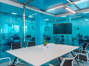 Modern glass meeting room with blue-tinted walls, white table, black chairs, and wall-mounted screen in a tech office.