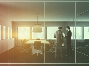 Two businessmen discuss documents inside a sleek glass meeting room with a large boardroom table and city views.