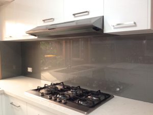Modern kitchen with stainless steel gas cooktop, white cabinetry, and a reflective white frosted glass splashback under rangehood.