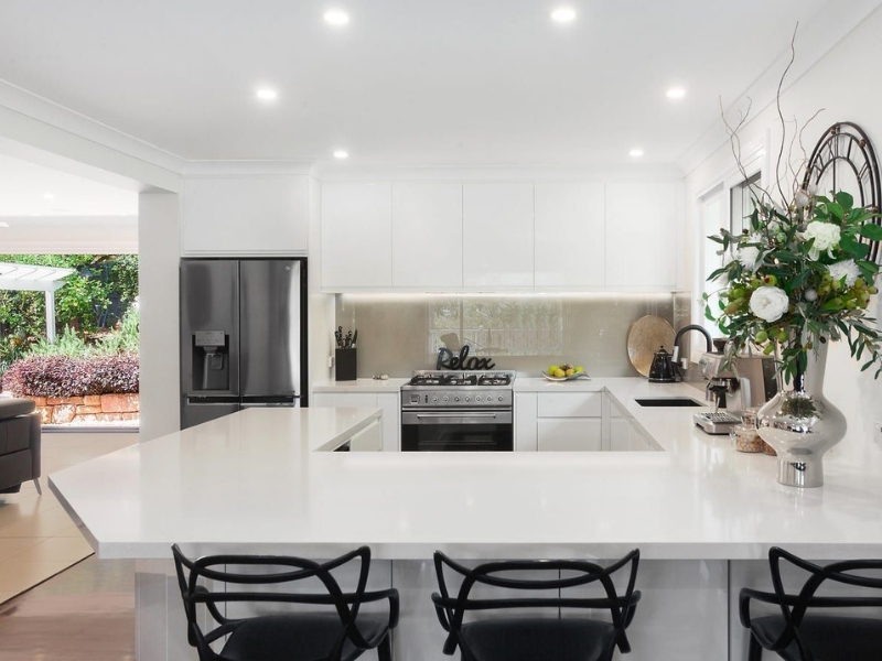 Bright open-plan kitchen with large white benchtop, black stools and a glossy white frosted glass splashback behind the stove.