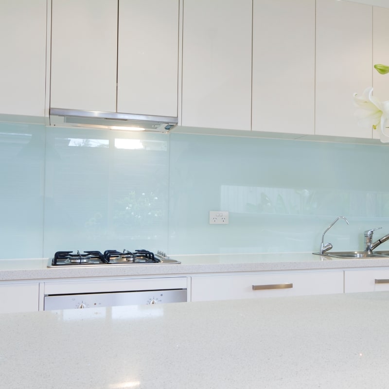 Kitchen splashback made of pale blue glass behind a gas cooktop and sink in a modern white kitchen